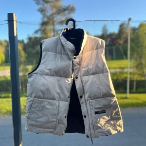 Beige canada goose väst - Säljer en beige dunväst från Canada Goose med svart insida och ståkrage. Västen har knappar framtill, två stora fickor och en diskret logotyp på bröstet. Perfekt för lager-på-lager under kyliga dagar.