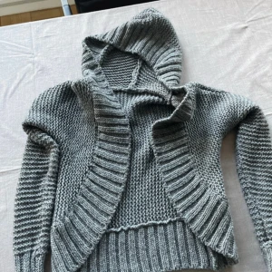 Grå stickad kofta med luva - Mysig grå stickad kofta med luva och breda ribbade kanter. Den är öppen fram och du kan välja hur du vill ha dina ärmar.