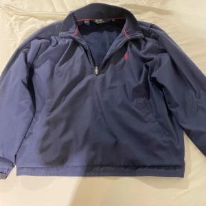 Blå halvzip jacka från Polo Ralph Lauren - Mörkblå jacka med halv dragkedja och röd broderad logga på bröstet. Klassisk krage och diskreta röda detaljer på insidan av kragen. Perfekt för en stilren och sportig look.
