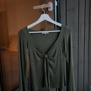DesignBy Si topp i militärgrönt - Säljer min fina topp från DesignBy Si som tyvärr inte kommer till användning.  Storlek XS/S, tröjan är normal i storleken och är endast testad. Den är lite skrynklig då den legat i en flyttkartong men det åtgärdas lätt med att stryka den. Nypris: 393 kr