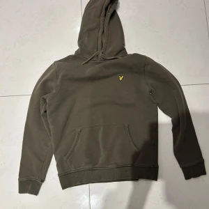 Olivgrön hoodie från Lyle & Scott - Säljer en olivgrön hoodie från Lyle & Scott med klassisk gul logga på bröstet. Tröjan har huva med dragsko och en stor magficka framtill. Perfekt för en avslappnad stil.