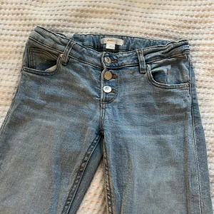 Ljusblå jeansbyxor med knappar - Säljer ett par ljusblå jeansbyxor med låg midja (bootcut). Byxorna har klassiska fem fickor och stängs med tre synliga knappar framtill. Perfekta till vardags och lätta att matcha med olika toppar. Säljer pågrund av att de har blivit för små.
