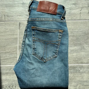 Tiger of Sweden jeans, modell Kelly - Snygga blå jeans från Tiger of Sweden i modellen Kelly. Klassisk femficksdesign med raka ben och bruna sömmar. Tillverkade i Italien och har en stilren läderpatch bak i midjan. Inga defekter