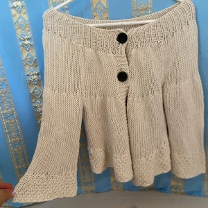 Beige stickad kofta med knappar från Åhléns - Säljer en beige stickad kofta från Åhléns med två stora svarta knappar framtill. Den har en lätt utställd form och bred ribbad midja. Perfekt för dig som gillar en mysig och avslappnad stil.