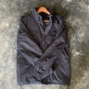 Mörkblå fieldjacka - Säljer en mörkblå fieldjacka i storlek medium. Jackan har dragkedja och tryckknappar framtill, hög krage och flera praktiska fickor. Perfekt för blåsiga dagar och enkel att matcha med olika outfits.