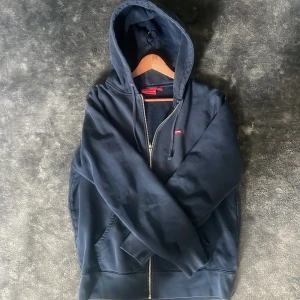Svart hoodie med dragkedja från Supreme - Säljer en svart hoodie från Supreme med dragkedja framtill och justerbar huva med snören. Klassisk Supreme-logga på bröstet och mjukt innerfoder. Perfekt för en avslappnad streetwear-look.