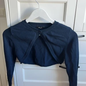Mörkblå croppad kofta från Gina Tricot - Säljer en mörkblå croppad kofta från Gina Tricot. Tröjan är stickad med lång ärm och har en knapp framtill vid halsen. Perfekt att slänga över en topp för en trendig lager-på-lager-look.