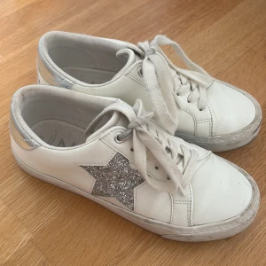 Vita sneakers med glittrig stjärna från Next - Säljer ett par vita sneakers från Next med en glittrig silverstjärna på sidan och silvriga detaljer vid hälen. Skorna har snörning och platt sula, perfekta för en trendig vardagslook. Ganska använda men inget som är sönder 🤗