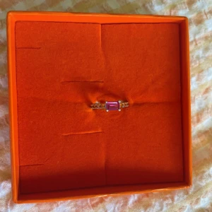 Izabel Display ring - Cool silvrig lillfinger ring ifrån Izabel Display. Den har orangea stenar runtom och en större rosa sten. Den är ca 15 mm i diameter vilket motsvarar storlek 46, och den är använd fåtal gånger och inga tecken på användning. Nypris: 800kr