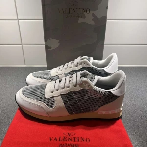 Valentino Garavani sneakers i grått och vitt - Snygga sneakers från Valentino Garavani i grått, vitt och svart med meshdetaljer och klassisk snörning. Skorna har en sportig design med rund tå och platt sula samt ikoniska nitar på hälen. Perfekta för dig som gillar exklusiva märken och stilren look.