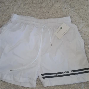 Lacoste Badbyxor - Snygga vita shorts från Lacoste med elastisk midja och snörning. Shortsen har två svarta ränder och Lacoste-logga framtill samt en bakficka. Perfekta för sommarens aktiviteter.