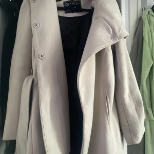 Beige kappa med bälte från New yorker  - Snygg beige kappa från New yorker med bred krage och knappar framtill. Kappan har ett matchande bälte i midjan och är helfodrad. Perfekt för kyligare dagar och ger en stilren look. Priset kan diskuteras! 