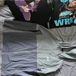 Grå vindjacka från Under Armour - Säljer en snygg tvåfärgad grå vindjacka från Under Armour med huva och dragkedja. Jackan har lång ärm, meshfoder på insidan och logga på bröstet. Perfekt för träning eller när det blåser ute.