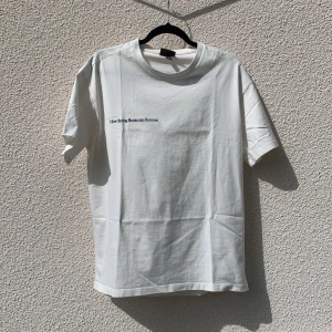 Vit t-shirt med tryck från H&M - Säljer en vit t-shirt från H&M med blå texten 'How Spring Bursts into Summer' på bröstet och ett stort fototryck av ett blomsterfält på ryggen. T-shirten är i 100% bomull och har en lätt oversized passform.