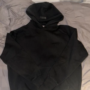 Svart hoodie från Essentials Fear of God - Säljer en svart hoodie från Essentials Fear of God med diskret logga på bröstet och stor logga på ryggen. Tröjan har huva och en klassisk känguruficka framtill. Perfekt för dig som gillar streetwear och enkel stil.