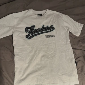 Vit t-shirt från Hoodrich med tryck - Vit t-shirt från Hoodrich med stort broderat Hoodrich-tryck på bröstet och tryck på ryggen. Klassisk passform och rund hals. Perfekt för dig som gillar streetwear och vill sticka ut med en snygg logga.