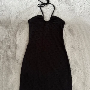 Svart halterneck klänning - Säljer en svart halterneck klänning med åtsittande passform och diskret strukturmönster. Klänningen har knyt runt nacken och är ärmlös, perfekt för en trendig look.