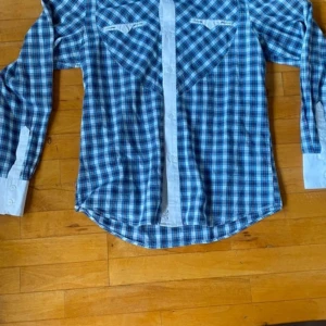 Blårutig skjorta från Armani Exchange - Säljer en blårutig skjorta från Armani Exchange i storlek L. Skjortan har långa ärmar, vita detaljer vid manschetter och knappslå samt klassisk krage. Perfekt för dig som gillar stilren och tidlös design.