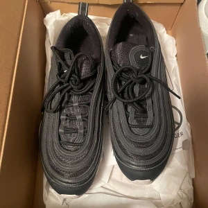 Svarta Nike Air Max 97 sneakers - Säljer ett par svarta Nike Air Max 97 sneakers med vit sula och klassisk vågig design. Skorna har snörning och en liten vit Nike-logga på sidan. Perfekta för dig som gillar en sportig och stilren look.