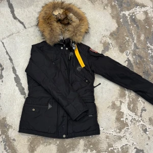 Svart vinterjacka med päls från Parajumpers - Säljer en svart vinterjacka från Parajumpers med äkta päls runt huvan och snygga detaljer som gul rem och broderad patch på ärmen. Jackan har dragkedja, knappar och flera fickor. Perfekt för kalla dagar.
