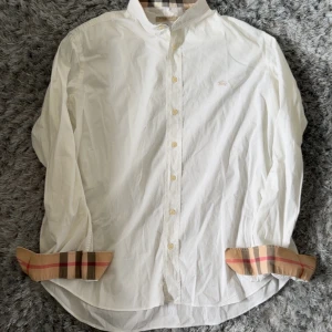 Vit skjorta från Burberry Brit med rutiga detaljer - Säljer en klassisk vit skjorta från Burberry Brit med ikoniska beige, svarta och röda rutiga detaljer på krage och ärmslut. Skjortan har knappar framtill och är långärmad. Perfekt för dig som gillar stilrena plagg med en twist.