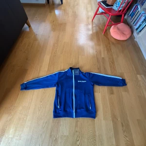 Palm Angels kofta - Snygg blå track jacket från Palm Angels med vit dragkedja och vita ränder längs ärmarna. Jackan har ståkrage, två fickor med dragkedja och logga på bröstet. Perfekt för en sportig och trendig look.