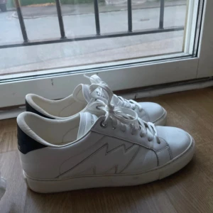 Vita sneakers från Zadig & Voltaire - Säljer ett par vita sneakers från Zadig & Voltaire med svart hälparti och vit sula. Skorna har snörning och en blixtdetalj på sidan. Perfekta nu inför sommaren🥰