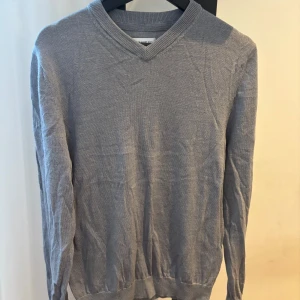 Grå v-ringad tröja från These Glory Days - Säljer en stilren grå v-ringad tröja från These Glory Days. Tröjan är tunnstickad med långa ärmar och ribbade muddar vid ärmslut och nederkant. Perfekt för lager-på-lager och enkel att matcha med det mesta.