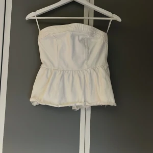 Vit tubtopp med volang - Säljer en vit tubtopp i denim från H&m med volang nedtill och slitna kanter. Toppen har dragkedja bak för enkel påklädning och är perfekt för sommardagar. Enkel och stilren design som passar till mycket. Endast använd en gång. Säljer pga för liten