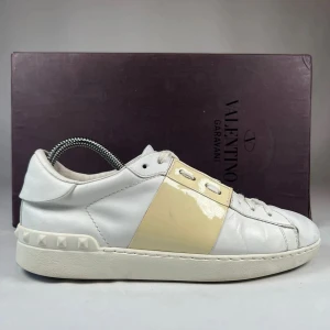 Valentino Open sneakers i vitt och beige - Säljer ett par snygga Valentino Open sneakers i vitt skinn med en beige lackad detalj på sidan. Skorna har rund tå, platt sula och dekorativa nitar på hälen. Perfekta för dig som gillar exklusiva och stilrena sneakers.