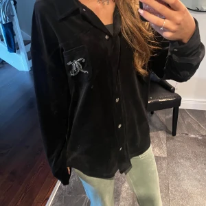 Svart skjorta från Juicy Couture - Säljer en svart skjorta från Juicy Couture med glittrig logga på bröstfickan och stor text på ryggen. Skjortan har klassisk krage, knäppning framtill och är tillverkad i ett mjukt, sammetsliknande material. Perfekt för dig som gillar statement-plagg!