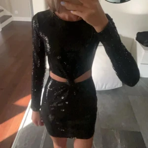 Svart glittrig kortklänning med cut-out - Säljer en svart glittrig kortklänning med lång ärm och snygg cut-out detalj i midjan. Klänningen har en tight passform och är perfekt för dig som vill sticka ut på festen. Materialet är glittrigt och skimrar snyggt i ljuset.