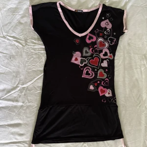 Svart kortklänning med hjärtmönster och rosa detaljer - Vintage y2k Svart kortklänning miniii eller tunica/long topp med ljusrosa kantband. Framsidan har tryckta hjärtan i rosa, rött och vitt. Klänningen har korta ärmar och är tillverkad i ett mjukt, stretchigt material. Öppen bak i ryggen. 