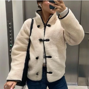 Beige teddyjacka med svarta detaljer - Säljer en beige teddyjacka med svarta knappar och kantdetaljer. Jackan har hög krage och lång ärm, samt en oversized passform. Perfekt för kyliga dagar och ger en trendig look.
