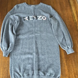 Grå stickad klänning från Kenzo - Snygg grå stickad klänning från Kenzo med broderad logotyp framtill i svart, vitt och ljusrosa. Klänningen har rund halsringning och långa, puffiga ärmar med ribbade muddar. Perfekt för en trendig och avslappnad look.