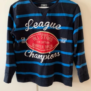 Mörkblå randig långärmad tröja med tryck - Säljer en mörkblå långärmad tröja från PRIMARK med blå ränder och stort tryck framtill med texten 'League Champions' och en röd amerikansk fotboll. Perfekt för sportiga dagar eller chill.