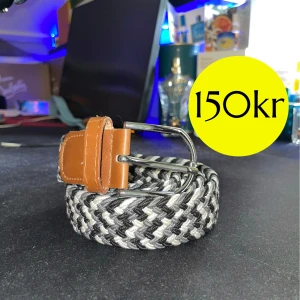 Svart vit flätat bälte - Snyggt flätat bälte i svart, vitt och grått med bruna skinn-detaljer vid spännet och änden. Bältet har ett klassiskt silverfärgat spänne och passar perfekt till jeans eller chinos. Ger en stilren och modern look.