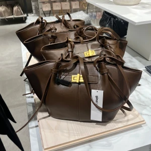 Brun axelväska med gulddetalj - Snygg brun axelväska i skinnimitation från Gina Tricot. Väskan har långa handtag, dekorativt spänne i guld och stilrena linjer. Perfekt för att bära dina viktigaste saker med stil. Den är endast använd fåtal ggr så skicket är därmed så gott som ny🪽