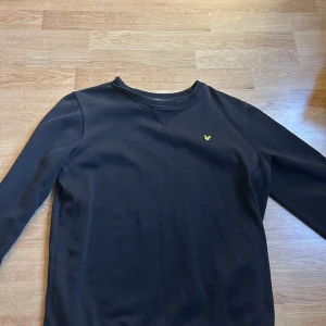 Svart sweatshirt från Lyle & Scott - Säljer en svart sweatshirt från Lyle & Scott Junior med klassisk rund halsringning och broderad gul logga på bröstet. Tröjan har långa ärmar och är tillverkad i mjuk bomull. Perfekt för en stilren och enkel look.