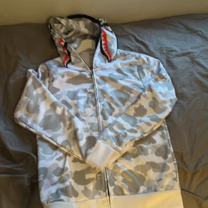 Vit och grå camouflage hoodie med hajmotiv - Säljer en hoodie från BAPE i vit och grå camouflage med dragkedja hela vägen upp över huvan. Huvan har ett hajmotiv med tänder och ögon i rött och svart. Perfekt för dig som gillar streetwear och unika plagg.