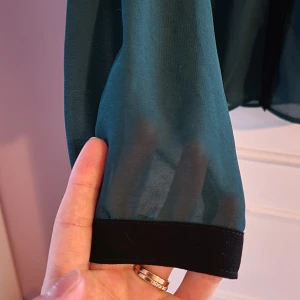 Mörkgrön transparent blus - Säljer en mörkgrön blus med svarta detaljer som är ganska transparent, se bild.