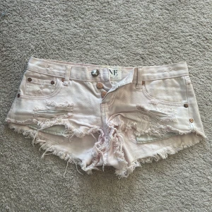 Jeansshorts One Teaspoon  - Säljer ett par ljusrosa jeansshorts med slitna detaljer och fransig kant. Shortsen har låg midja, klassiska fickor och knappar framtill. Perfekta för sommardagar och en avslappnad stil. Storlek W26