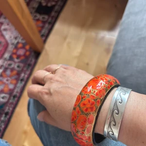 Trädarmband med blommönster - Säljer ett färgstarkt och brett armband i orange med röda och guldiga blommor. Armbandet har ett unikt och iögonfallande mönster som verkligen sticker ut. Perfekt för dig som vill addera lite färg till din stil! Handmålad 