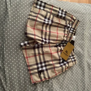 Rutigta badshorts från Burberry - Snygga badshorts från Burberry i klassiskt beige rutigt mönster med svarta, vita och röda ränder. Shortsen har elastisk midja och Burberry-logga broderad på benet. Perfekta för sommaren!