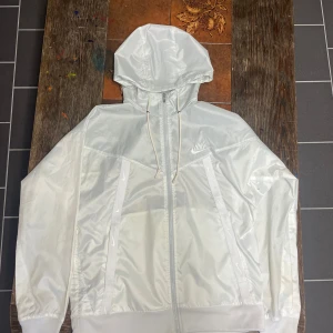 Vit vindjacka från Nike med huva - Säljer en stilren vit vindjacka från Nike i storlek M. Jackan har huva, dragkedja framtill och två snedställda fickor. Tillverkad i lätt syntetmaterial, perfekt för blåsiga dagar. Klassisk Nike-logga på bröstet och justerbar huva med snören. DOCK har den fått lite grön sprayfärg på framsidan och så har den lite gul missfärgning på baksidan. Men den gula missfärgningen märks knappast. Kan bytas mot något likvärdigt i storlek M/L. Hämtas i Skövde annars fraktas det.