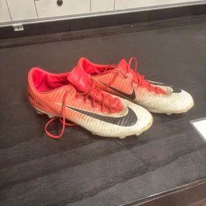Nike Mercurial fotbollsskor i rött och vitt - Säljer ett par Nike Mercurial fotbollsskor i rött och vitt med svarta detaljer. Skorna har snörning och är designade för att ge bra grepp på planen. Perfekta för dig som vill ha lätta och snygga skor till fotbollsträningen.