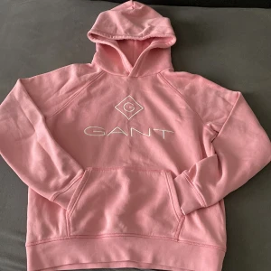 Rosa hoodie från GANT - Säljer en ljusrosa hoodie från GANT med broderad logga framtill. Tröjan har huva och en stor magficka. Perfekt för dig som gillar klassisk och stilren streetwear.
