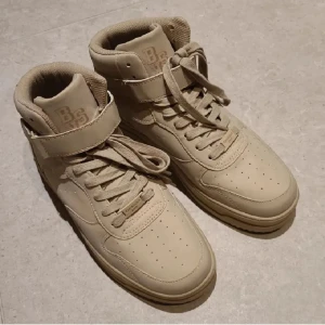 Beiga sneakers från Bershka - Snygga beige sneakers från Bershka med hög modell, snörning och band i stlk 41. Skorna har en bred rem över vristen för extra stil och komfort. Perfekta för en trendig look. Endast testade.