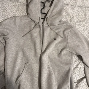 Ralph lauren hoodie Grå - Säljer en stilren ljusgrå hoodie från Polo Ralph Lauren med dragkedja och huva. Tröjan har två fickor framtill och en broderad logga på bröstet. Perfekt för en avslappnad look. Köpt för 1900 på zalando mitt pris är 850