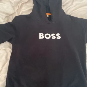 Svart hoodie från BOSS - Säljer en svart hoodie från BOSS med vit logga tryckt på bröstet. Tröjan har huva och är tillverkad i bomull. Perfekt för en avslappnad och stilren look.